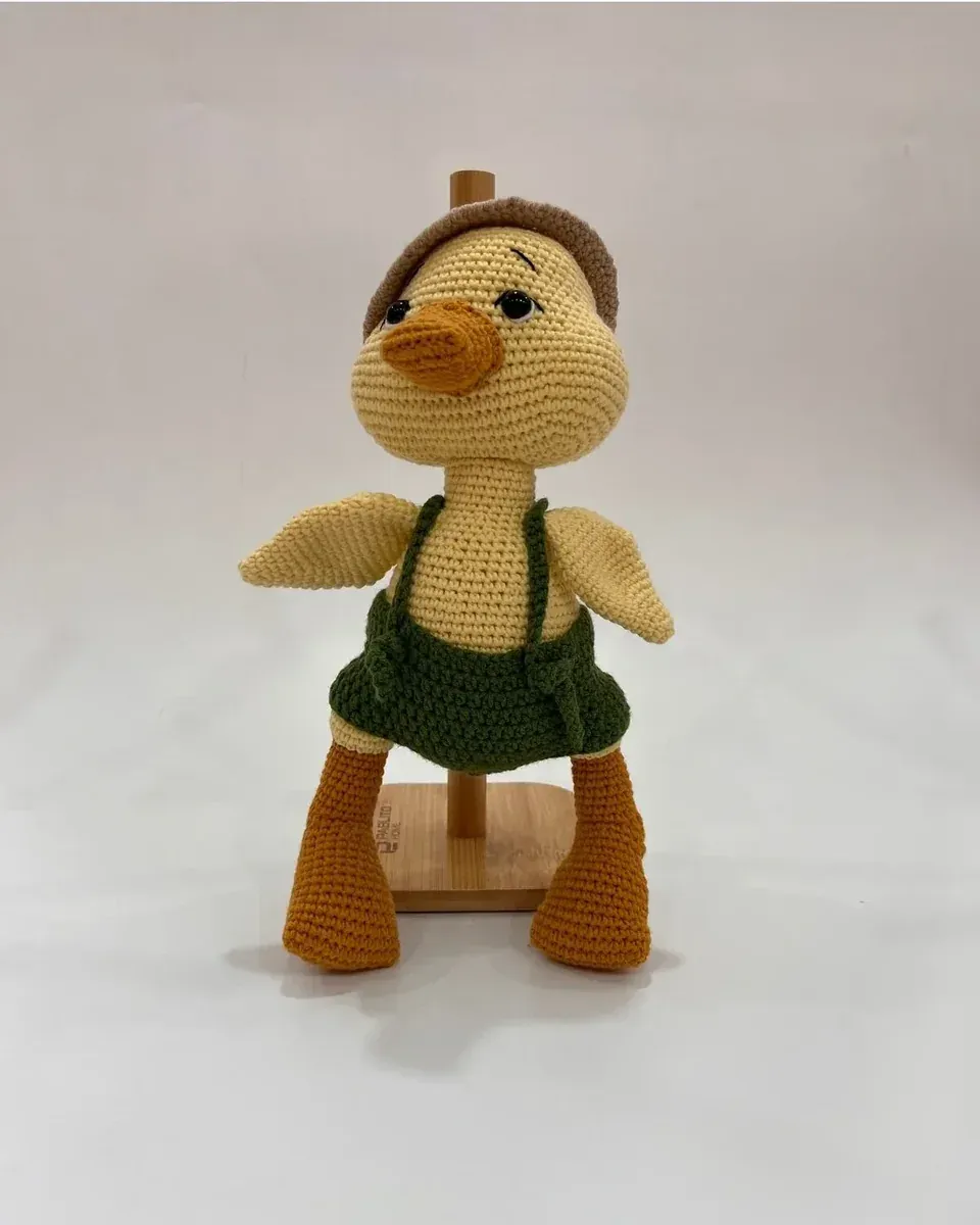 Yeşil salopetli sarı amigurumi ördek, ahşap stant üzerinde