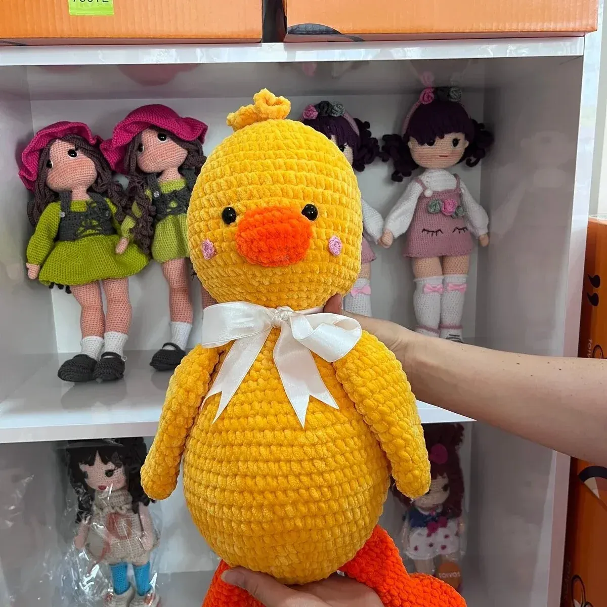 Mağaza rafındaki amigurumi bebeklerin önünde elde tutulan fiyonklu sarı ördek